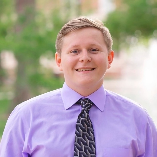 A young man with light brown hair and blue eyes smiles broadly, wearing a light purple collared shirt and a dark geometric patterned tie against a blurred green background.