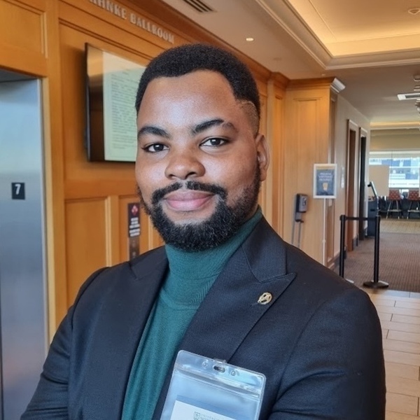 A Black man with a beard and a slight smile wears a dark suit jacket, green turtleneck, and an ID badge. He stands near a wood-paneled elevator and a digital sign for the Ranke Ballroom at Notre Dame.