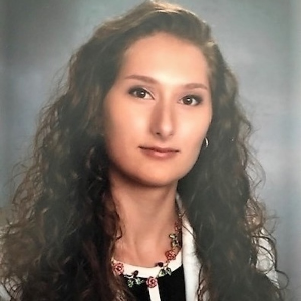 A young woman with long, dark curly hair and a neutral expression looks forward. She wears a black top, white blazer, and a necklace with pink roses and green leaves against a light blue-grey background.