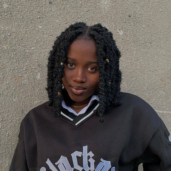 Young Black woman with shoulder-length black curly braids, some adorned with gold beads, smiles gently. She wears a black V-neck sweater with "black" text over a white striped-collar shirt, against a grey wall.