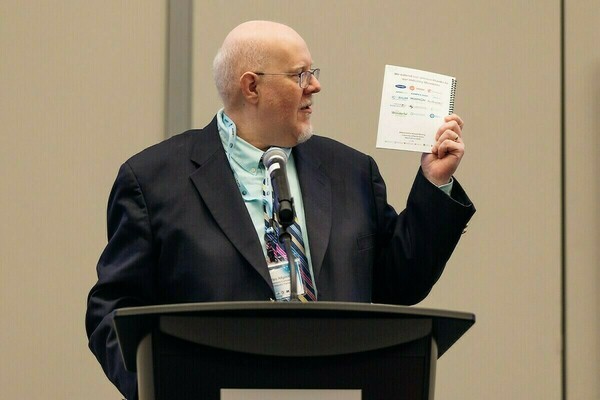A man with glasses and white hair, wearing a dark suit, light blue shirt, and striped tie, speaks at a podium. He holds up a spiral-bound booklet from the "PRISM-ERA Annual Meeting University of Notre Dame" that thanks industry members.