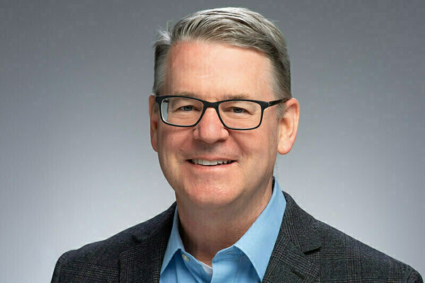 A man with grey hair and black-framed glasses smiles, wearing a light blue collared shirt and a dark grey plaid blazer.