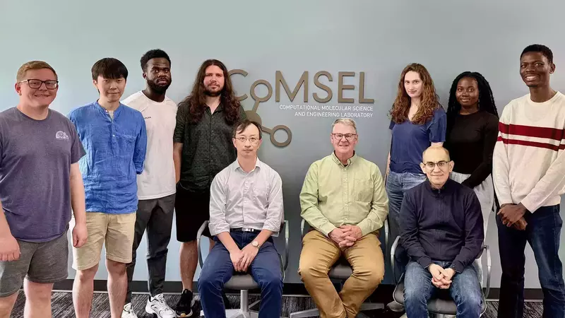 Ten people, seven standing and three sitting, smile for a group photo in front of a blue wall with the COMSEL lab logo. They wear a mix of casual shirts in colors like blue, white, green, and black.